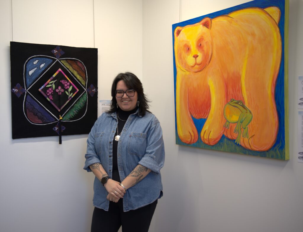 Shay Paul stands between two of Stínesten Gallery's art pieces, smiling. (Photo Taken by ___, TRU, 2026)